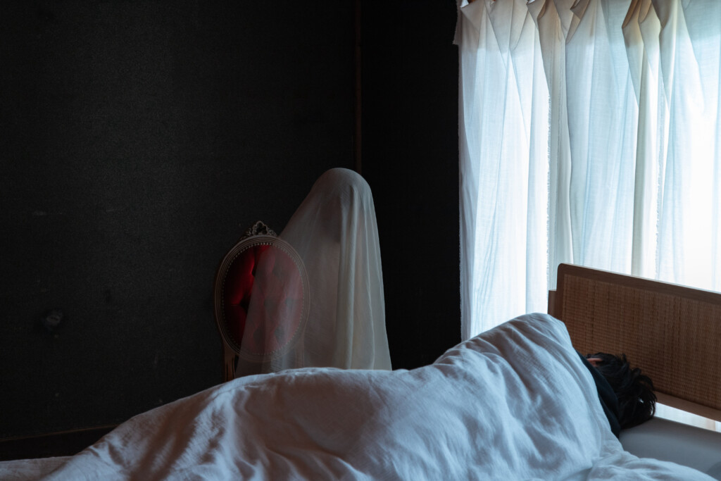 ベッドで寝込んでいる男性を心配そうに看病する白い布を纏ったオバケのフォトコラージュ作品。