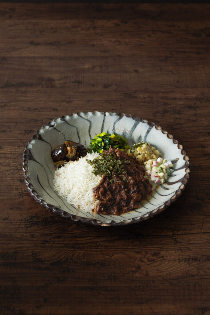 野菜や肉の持つ水分だけで鶏肉が柔らかくなるまで煮込まれた具材とスパイスが絶妙に溶け合い、うまみが凝縮した無水チキンカリー。ライスとカレーの周りの副菜を混ぜることで味の変化も楽しめる。